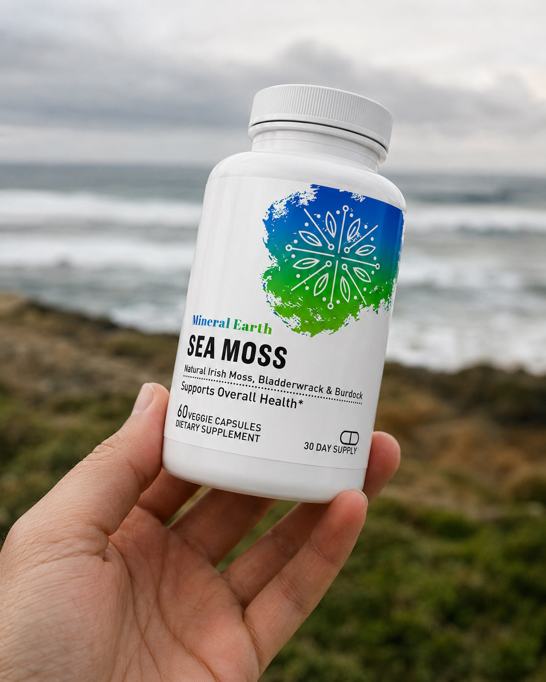 Hand holding Mineral Earth Sea Moss supplement bottle on rocky ocean shore